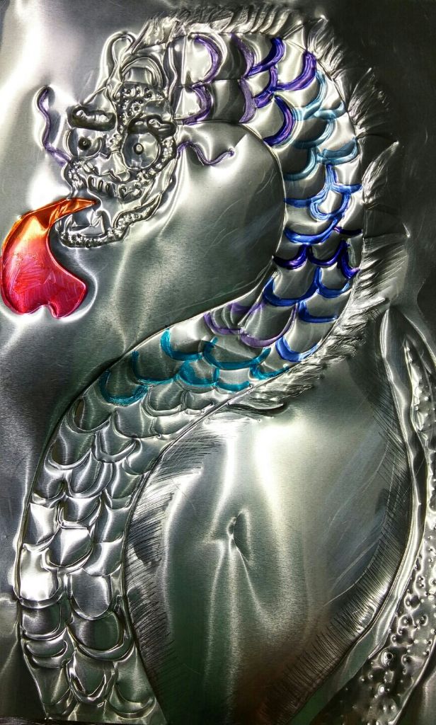 A dragon worked in relief on a metal sheet, with raised scales and a red-orange flame issuing from its mouth. The scales are inked in blue, purple, and teal.